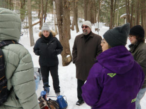 A small group of people in winter coats and toques chatting in a snowy forest, with blue enamel coffee pots nearby