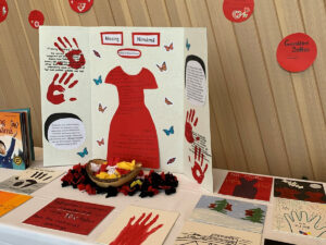 A display table with a red dress cut-out, handprints and drawings honouring missing and murdered Indigenous women, with books and a bowl of red ribbons