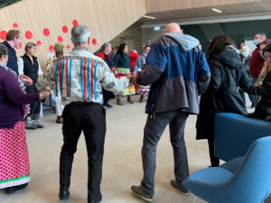 People gathered in a circle in a bright hall decorated with red circles, some in traditional ribbon skirts and shirts