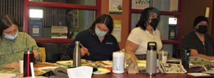 Four masked people painting at a table in a workshop room