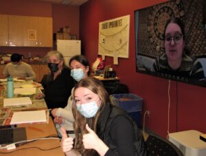 Participants seated at a table in a painting workshop, one giving a thumbs up, with a remote participant on a screen on the wall