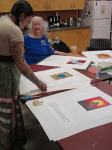 A woman arranging printed artworks on a table while an older woman looks on