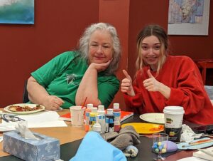 Two smiling women giving thumbs up at a table with paint bottles, a coffee cup and a tissue box