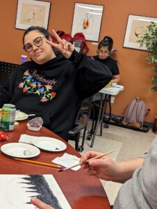 A woman in a floral-embroidered hoodie making a peace sign at a table during a painting workshop