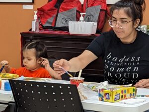 A woman and a toddler painting at a table with craft supplies, the toddler in an orange shirt