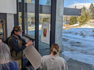A man in a vest and cap holds a large sheet of paper up to a glass wall inside a building, with snow outside