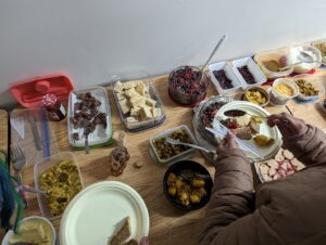 Overhead view of a table with a potluck of traditional foods in containers, and a person serving a plate