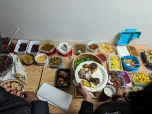 Overhead view of a table with a large potluck of dishes in containers and a person holding a loaded plate