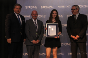 Dave Marshall Leadership Awards ceremony photo of an award recipient holding a framed certificate in front of a Nipissing University backdrop