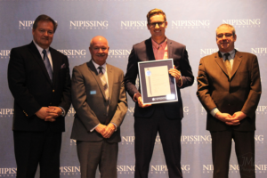 Dave Marshall Leadership Awards ceremony photo of an award recipient holding a framed certificate in front of a Nipissing University backdrop