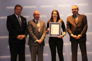 Dave Marshall Leadership Awards ceremony photo of an award recipient holding a framed certificate in front of a Nipissing University backdrop