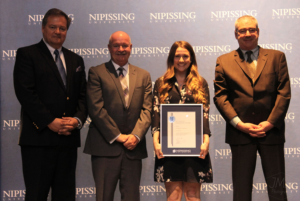 Dave Marshall Leadership Awards ceremony photo of an award recipient holding a framed certificate in front of a Nipissing University backdrop