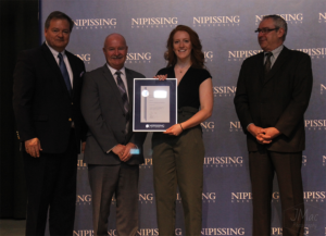Dave Marshall Leadership Awards ceremony photo of an award recipient holding a framed certificate in front of a Nipissing University backdrop
