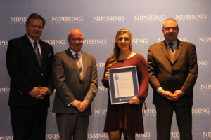 Dave Marshall Leadership Awards ceremony photo of an award recipient holding a framed certificate in front of a Nipissing University backdrop