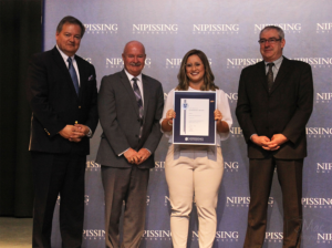 Dave Marshall Leadership Awards ceremony photo of an award recipient holding a framed certificate in front of a Nipissing University backdrop