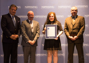 Dave Marshall Leadership Awards ceremony photo of an award recipient holding a framed certificate in front of a Nipissing University backdrop