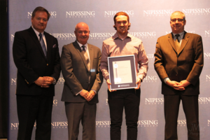 Dave Marshall Leadership Awards ceremony photo of an award recipient holding a framed certificate in front of a Nipissing University backdrop