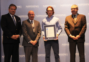 Dave Marshall Leadership Awards ceremony photo of an award recipient holding a framed certificate in front of a Nipissing University backdrop