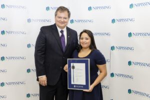 A man in a dark suit stands next to a woman holding a framed Dave Marshall Leadership Award certificate in front of a Nipissing University backdrop