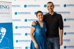 A smiling man and woman posing with their arms around each other in front of a Nipissing University backdrop, next to a Dave Marshall Awards banner