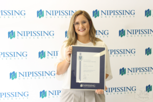 A smiling woman with glasses holds a framed Dave Marshall Leadership Award certificate in front of a Nipissing University backdrop